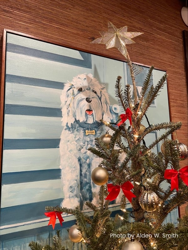 A painting of a shaggy white dog hangs on a wooden wall behind a Christmas Tree