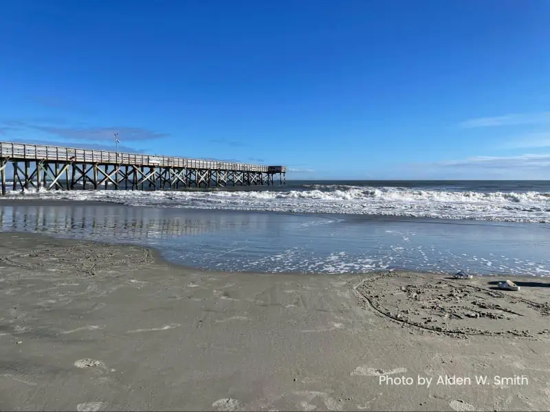 A wooden pier extends into a blue ocean with a modicum of surf in the foreground.