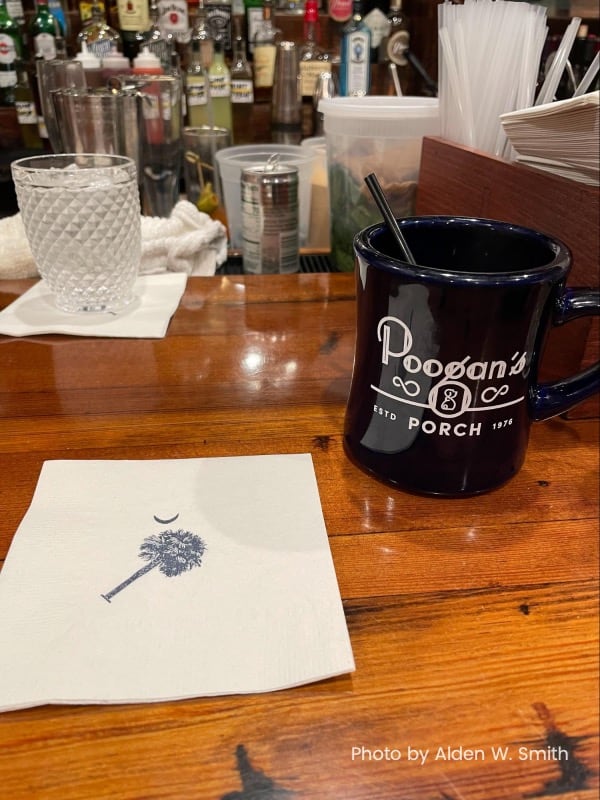 A blue coffee cup sits on a polished wooden bar next to a white cocktail napkin with the state logo for South Carolina on it.  A full bar rounds out the background.