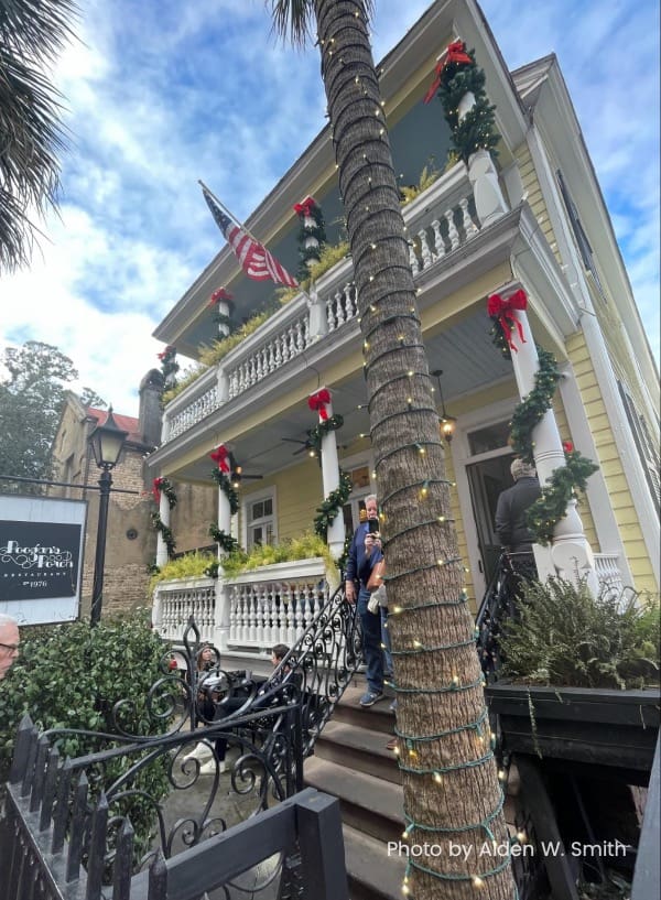 A yellow, three-story house with a white, terraced porch and black, wrought-iron fence. Palm trees decorate the foreground.
