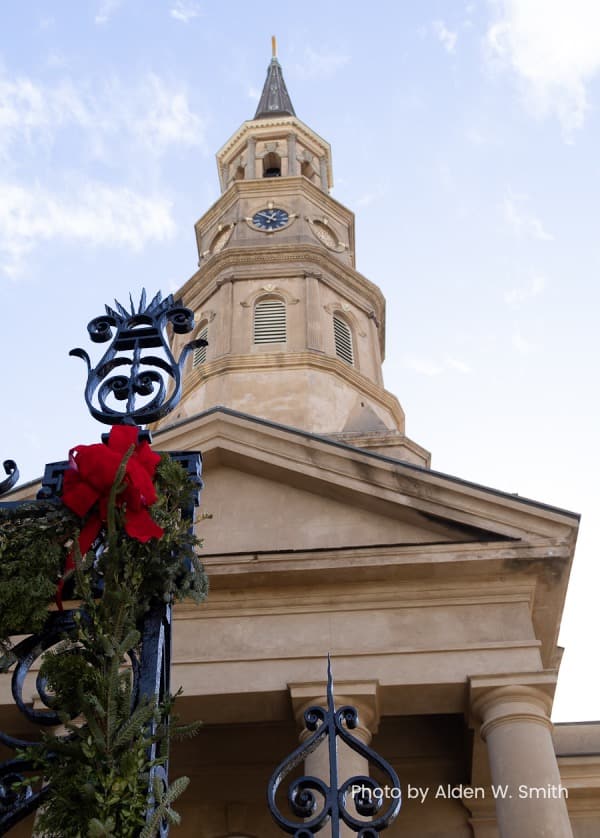 A tall, stone, church steeple rises into a blue sky.  Holiday wreathes cover the wrought-iron fence in the foreground